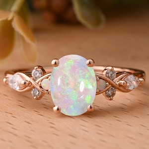 May include: Rose gold ring with an oval opal centre stone displaying iridescent colours. The ring is embellished with small, clear gemstones and decorative flourishes on the band. A beautiful piece of jewellery.