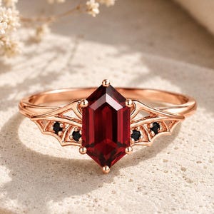 Puede incluir: Un anillo de oro rosa con una piedra preciosa de color rojo intenso y corte hexagonal en el centro. La banda presenta un intrincado diseño de metal en forma de alas a ambos lados, con pequeñas piedras negras redondas incrustadas en la estructura.
