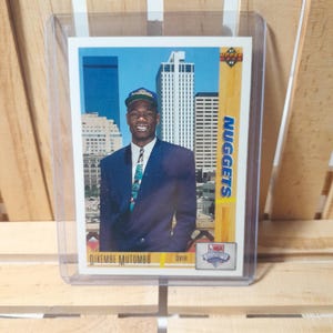Può includere: Una carta da collezione di basket con Dikembe Mutombo, un centro dei Denver Nuggets. La carta è della serie Upper Deck NBA Hoops 1995-96. La carta presenta una foto di Mutombo con la sua divisa dei Nuggets, in piedi davanti a uno skyline cittadino. La carta include anche il nome di Mutombo, la sua posizione e il logo della squadra.