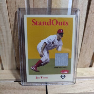 Puede incluir: Una tarjeta de béisbol con Jim Thome, un ex jugador de la Major League Baseball. La tarjeta es de la serie "StandOuts" y presenta una foto de Thome con un uniforme de los Philadelphia Phillies. La tarjeta también incluye un parche del jersey usado por Thome en un juego.