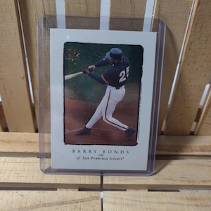 Puede incluir: Tarjeta de béisbol con Barry Bonds de los Gigantes de San Francisco en acción. La tarjeta muestra a Bonds con un uniforme negro y naranja, bateando. La tarjeta está dentro de una funda protectora de plástico.