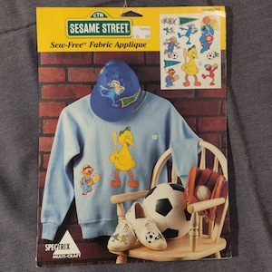 Op de afbeelding: Sew-Free Fabric Applique-set met Sesamstraat-personages. De set bevat een blauwe baseballpet met een Sesamstraat-personage, een blauw sweatshirt met een geel Big Bird-personage, een honkbalknuppel en een honkbal. De set bevat ook een vel met strijkbare applicaties met verschillende Sesamstraat-personages.