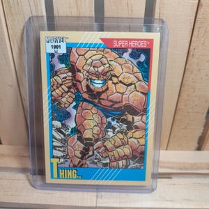 Op de afbeelding: Een Marvel Super Heroes trading card uit 1991 met The Thing. De kaart heeft een beige achtergrond met een blauwe en rode rand. The Thing wordt afgebeeld in een dynamische pose, met een gedetailleerd, rotsachtig oranje exterieur en een blauw kledingstuk.
