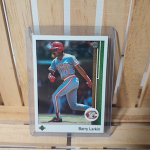 Puede incluir: Una tarjeta de béisbol con Barry Larkin con el uniforme de los Rojos de Cincinnati. La tarjeta muestra a Larkin en acción, con casco y guantes rojos, con el logo del equipo visible. La tarjeta está en una funda protectora.