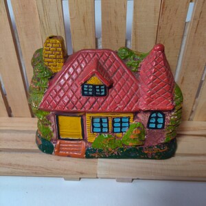 May include: A small ceramic house figurine with a red roof, yellow trim, and green accents. The house has a chimney, windows, and a door. The figurine is painted in a whimsical style.