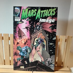 Puede incluir: Una portada de cómic con una ilustración verde, roja y negra de un personaje con una cabeza grande y un arma. El título del cómic es "Mars Attacks! image" y el número de la edición es 1.