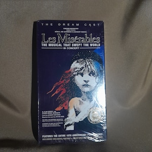 May include: A VHS tape featuring the musical Les Misérables, the musical that swept the world in concert. The cover features a black and white illustration of a young girl with red hair, against a blue and black background. The text "The Dream Cast" is at the top of the cover, and the text "Les Misérables" is in large, bold letters.