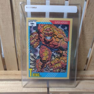 Op de afbeelding: Een Marvel Super Heroes-verzamelkaart uit 1991 met The Thing. De kaart toont een gedetailleerde illustratie van het personage in tinten oranje, bruin en blauw. De kaart zit in een beschermende plastic hoes met de tekst "BECKETT SHIELD".