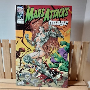Puede incluir: Una portada de cómic con una mujer de pelo rojo largo y un traje blanco y escamoso. Está de pie sobre un fondo rojo y naranja con otros personajes, incluyendo un extraterrestre verde y una figura con un traje negro y rojo. El título del cómic es "Mars Attacks!" y la editorial es "Image".