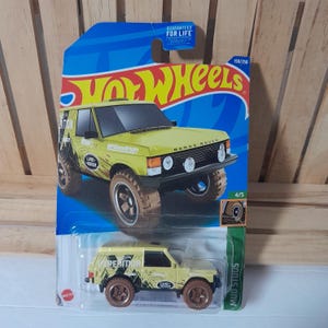 May include: A yellow Hot Wheels die-cast toy car of a Land Rover Range Rover. The toy car has brown wheels and black accents. The packaging is blue and features the Hot Wheels logo and the text "GUARANTEED FOR LIFE".