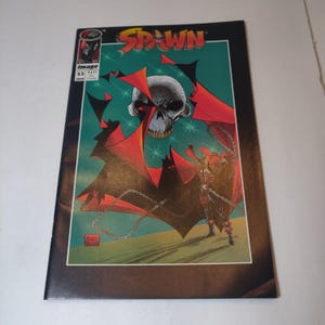 May include: A comic book cover featuring Spawn, a red-caped superhero with a skull face, standing on a chain-link fence. The cover is green and features a large skull with a red cape. The title "Spawn" is written in white letters on a black background.