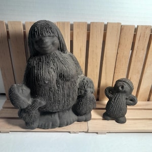 May include: Two dark gray, textured statues. One depicts a seated figure holding two smaller figures. The other is a small, standing figure with arms raised. The statues are on a wooden slatted surface.