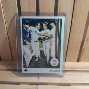 Puede incluir: Una tarjeta de béisbol con una foto de cuatro jugadores de béisbol con uniformes blancos y azules, celebrando en un campo de béisbol. La tarjeta está dentro de una funda de plástico transparente. La tarjeta dice "Kirk Gibson".