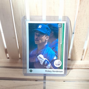 Puede incluir: Una tarjeta de béisbol con Rickey Henderson con el uniforme de los Yankees de Nueva York. La tarjeta tiene un borde blanco con detalles en verde y dorado. Henderson lleva una gorra azul y sostiene una pelota de béisbol. La tarjeta está en una funda protectora.