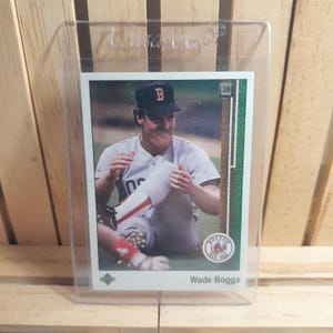 Puede incluir: Una tarjeta de béisbol con Wade Boggs con el uniforme de los Medias Rojas de Boston, dentro de un soporte de plástico transparente. La tarjeta tiene un borde verde y el logotipo de los Medias Rojas de Boston. El texto "Wade Boggs" está impreso en la parte inferior.