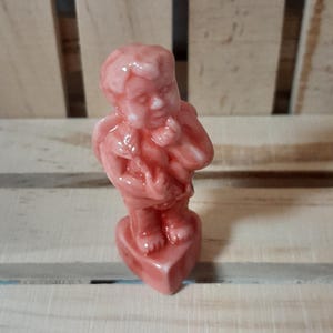 May include: A small, pink cherub figurine. The cherub is standing on a heart-shaped base. The figurine has a glossy finish and is made of ceramic or porcelain. The cherub is holding something in its arms.