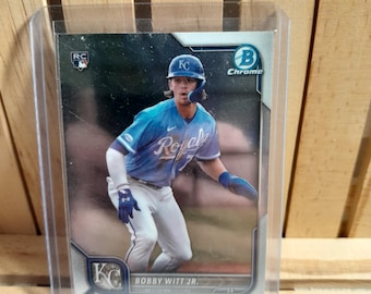 Peut inclure: Carte de baseball de Bobby Witt Jr. en uniforme des Royals de Kansas City, dans une pochette de protection. La carte porte le logo RC. Le joueur porte un uniforme bleu et blanc et un casque.