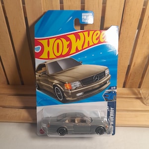 May include: A Hot Wheels die-cast model car in a package. The car is a gray coupe with black wheels and tan interior. The packaging is blue with the Hot Wheels logo and a picture of the car. The package reads "WIDEBODY".