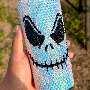 May include: A cylindrical tumbler fully covered in iridescent, light-reflecting rhinestones. The surface features a prominent, stylised face design created with black rhinestones, depicting two large, angular eyes and a wide, stitched-style mouth.