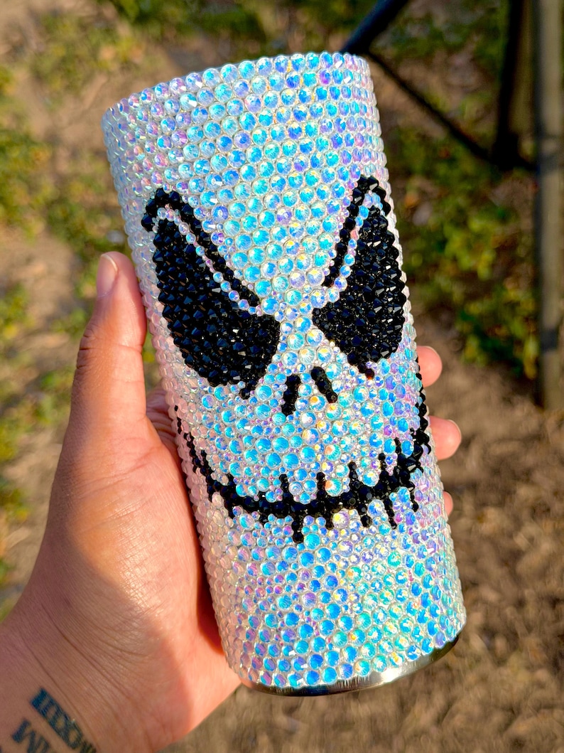 May include: A cylindrical tumbler fully covered in iridescent, light-reflecting rhinestones. The surface features a prominent, stylised face design created with black rhinestones, depicting two large, angular eyes and a wide, stitched-style mouth.