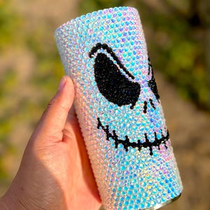 May include: A cylindrical tumbler fully covered in iridescent, light-reflecting crystals. The surface features a large, stylised face design created with black crystals, depicting two slanted, almond-shaped eyes and a wide, stitched-style mouth.