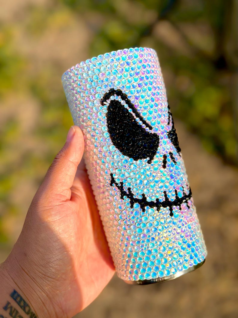 May include: A cylindrical tumbler fully covered in iridescent, light-reflecting crystals. The surface features a large, stylised face design created with black crystals, depicting two slanted, almond-shaped eyes and a wide, stitched-style mouth.