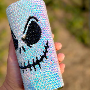 May include: A cylindrical tumbler fully covered in iridescent, light-reflecting rhinestones. The surface features a large, stylised face design created with black rhinestones, depicting two large oval eyes and a wide, stitched-style mouth.