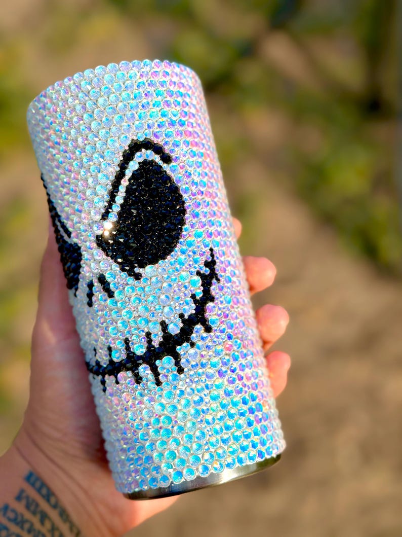 May include: A cylindrical tumbler fully covered in iridescent, light-reflecting rhinestones. The surface features a large, stylised face design created with black rhinestones, depicting two large oval eyes and a wide, stitched-style mouth.
