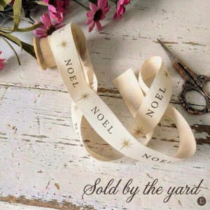 May include: Cream-colored ribbon with the word "NOEL" printed in black, along with starburst designs. The ribbon is spooled on a wooden spool. A pair of vintage scissors and pink flowers are in the background.