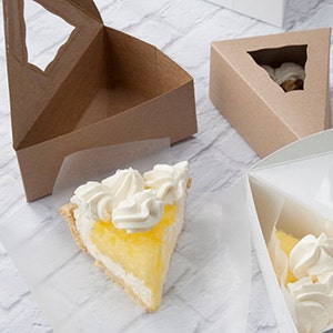 May include: A slice of lemon meringue pie with a golden crust and a creamy yellow filling, topped with swirls of white whipped cream. The pie sits on a white surface next to brown and white triangular pie boxes.
