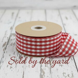May include: A spool of ribbon featuring a red and white gingham check pattern. The ribbon is wrapped around a circular cardboard core, with a portion of the fabric extending outward. The text "Sold by the yard" is printed in red script.