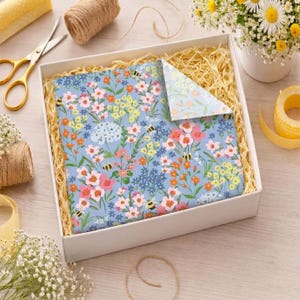 May include: A gift box with floral wrapping paper featuring pink, yellow, and orange flowers, and bees on a light blue background. The box is filled with straw and is surrounded by scissors, twine, and flowers.