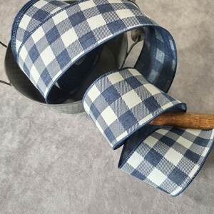 May include: A roll of blue and white checkered ribbon. The ribbon is a wide, decorative style with a gingham pattern. The ribbon is displayed in a metal bucket with a wooden handle. The ribbon is a perfect accent for crafts and gift wrapping.