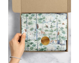May include: A box with a gold sticker on it. The box is filled with white tissue paper with a forest scene printed on it. The scene includes trees, birds, and animals.