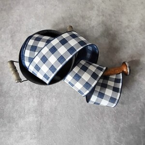 May include: A roll of blue and white checkered ribbon sits in a black metal container with wooden handles. A wooden dowel with a silver cap is used to hold the ribbon. The ribbon is a classic buffalo plaid pattern.