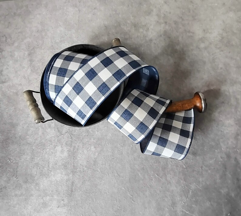 May include: A roll of blue and white checkered ribbon sits in a black metal container with wooden handles. A wooden dowel with a silver cap is used to hold the ribbon. The ribbon is a classic buffalo plaid pattern.