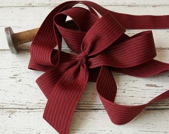 May include: Burgundy ribbon with thin black stripes, tied into a bow. The ribbon is wrapped around a wooden spool with a metal end. The ribbon is on a white, weathered wooden surface.