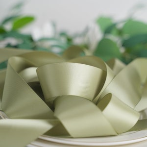 May include: A close-up of a sage green satin ribbon, twisted and folded, on a white plate. The ribbon is soft and smooth, with a subtle sheen.