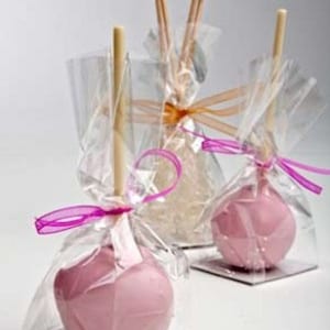 May include: Three pink cake pops on sticks, wrapped in clear cellophane bags with pink ribbon bows. The cake pops are decorated with a white swirl design.