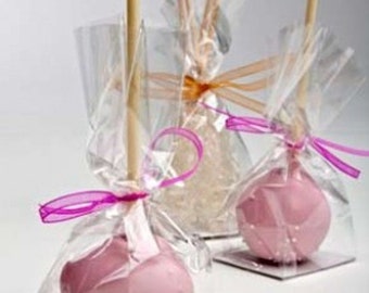 Puede incluir: Tres cake pops rosas en palitos, envueltos en bolsas de celofán transparentes con lazos de cinta rosa. Los cake pops están decorados con un diseño de remolino blanco.