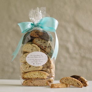 May include: A clear plastic bag filled with biscotti cookies tied with a light blue ribbon. The bag has a white label with the text "Fine Ingredients, Inc.  Hand-made Italian Style Biscotti.  P.O. Box 1391 - Mountainside, NJ 07092  (908) 541-1650  www.fineingred.com  Mixed Variety".  There are also several biscotti cookies on a white surface.