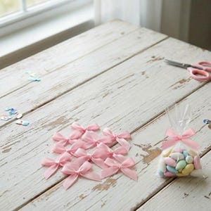 May include: A collection of pink satin bows and a small cellophane bag filled with pastel-colored candies. The bows are scattered on a distressed white wooden surface. Pink scissors are visible in the background, along with scattered confetti.