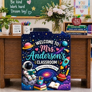 Op de afbeelding: Een rechthoekig klaslokaalbord met een donkerblauw ruimtethema met sterren, planeten, een raket en een lezende astronaut. De tekst luidt: Welcome to Mrs. Anderson's Classroom en Wheee Learning Takes Flight. Het bevat kleurrijke illustraties van boeken, een pennenhouder en een sterrenstelsel.