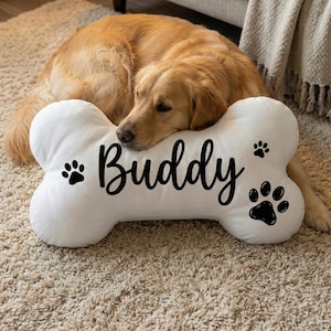 May include: A white, bone-shaped dog pillow with the name "Buddy" in black script. Black paw prints are on either side of the name. A golden retriever rests on the pillow, which is on a beige carpet.