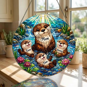 May include: Round stained glass window art featuring a family of otters in a river scene. The central otter has the word "Mom" on its chest, with three smaller otters labeled "Luna", "Benny", and "Finn". The background includes a waterfall, water lilies, and a sunburst.