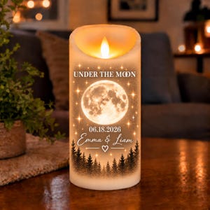 May include: A cylindrical LED pillar candle with a warm glowing flame. The exterior features a printed design of a full moon, a dark forest silhouette, and scattered stars. Text reads: UNDER THE MOON, 06.18.2026, and Emma & Liam with a heart.