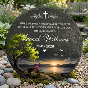 May include: A circular, dark grey memorial stone featuring a painted landscape. The scene depicts a lake, sunset, and a red bird on a bench. Text includes a cross, the name "Lional Williams", dates 1950-2026, and a quote.