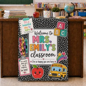 Op de afbeelding: Een zwart-wit patroon klaslokaal bord met de tekst Welcome to MRS. EMILY'S classroom, een gele schoolbus, een rode appel, kleurrijke kleurpotloden, alfabetblokken en een klembord met de tekst In a world where you can be anything be kind.