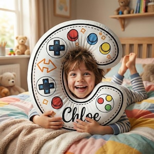 May include: A white, C-shaped pillow with a video game controller design. The pillow features red, blue, and yellow buttons, directional pads, and the name "Chloe" in black script. A child is smiling and lying on a bed with the pillow.