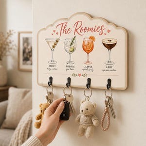 May include: A rectangular wooden wall-mounted key holder with a scalloped top edge, featuring the text The Roomies and illustrations of four cocktails labelled Emily dirty martini, Sophie gin tonic, Olivia aperol spritz, and Abby espresso martini.